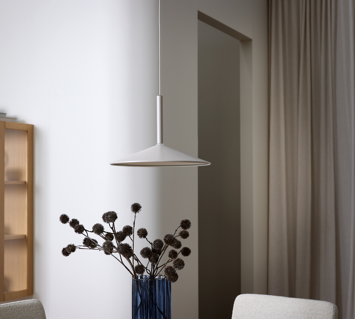 Hvid kegleformet pendel over blå vase med tørrede grene i minimalistisk stue med neutrale gardiner.