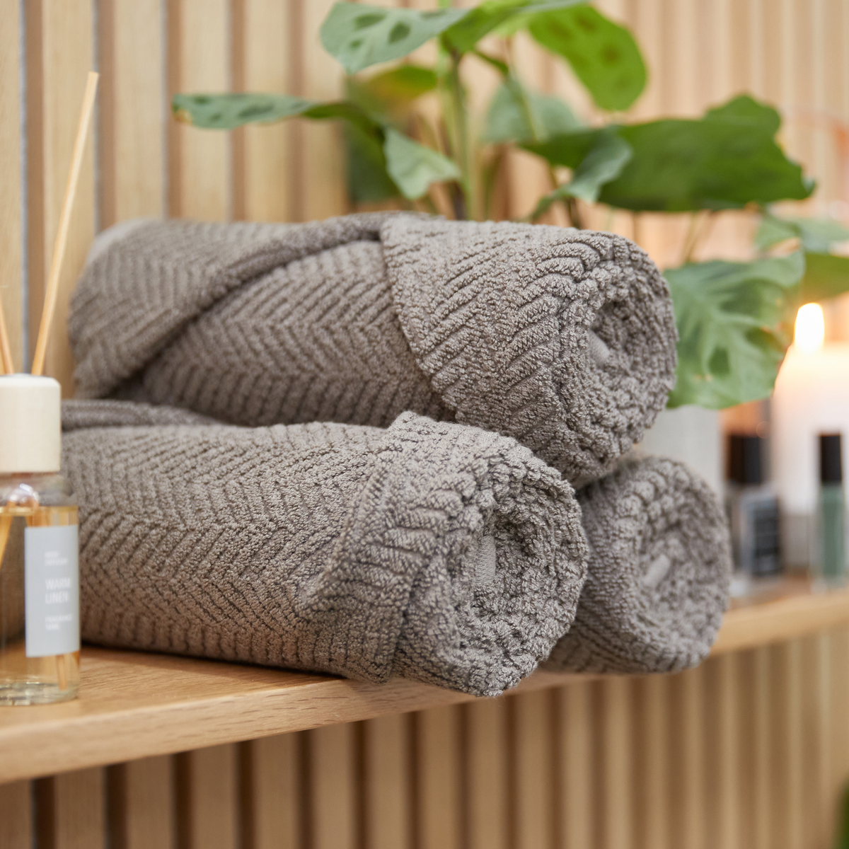 Tre rullede grå håndklæder på en træhylde med grøn plante, tændte lys og en duftpindflaske mærket ”WARM LINEN” i baggrunden.