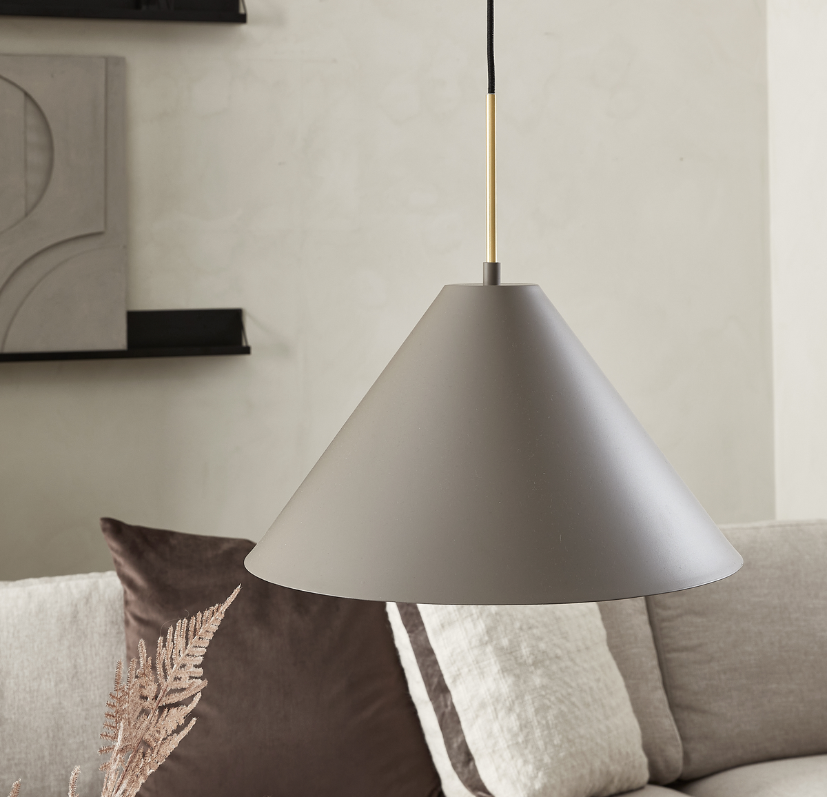 Konisk pendel i mat greige med messingsdetalje hænger over en beige sofa med puder i en minimalistisk stue.