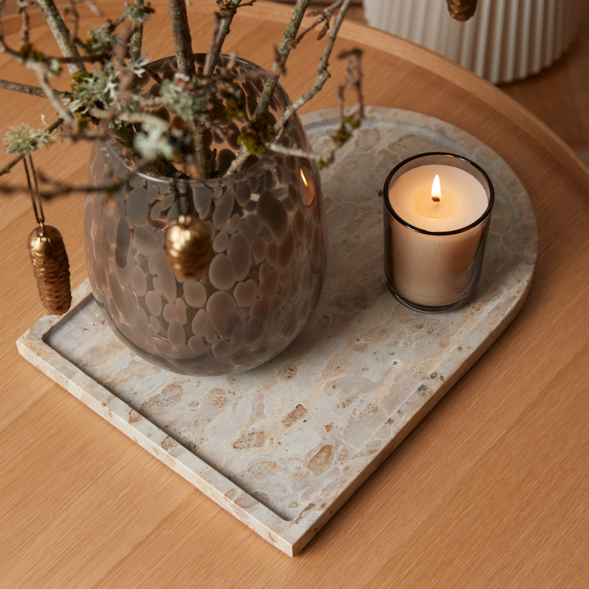 Røget glasvase med grene og tændt duftlys på beige stenbakke på et træbord.