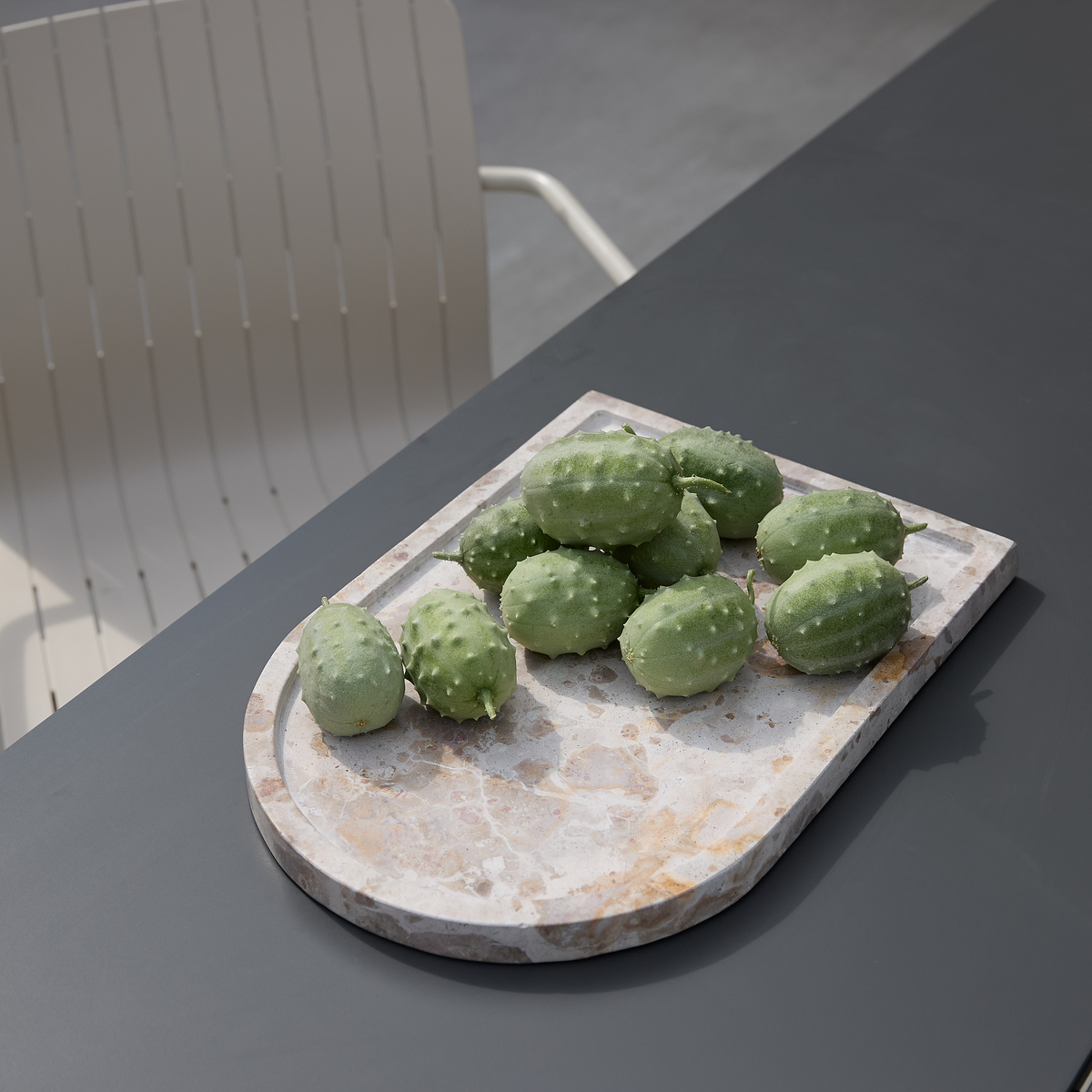 Grønne piggede frugter på et terrazzo-serveringsbræt på et mørkegråt udendørs bord med en lys stol i baggrunden.