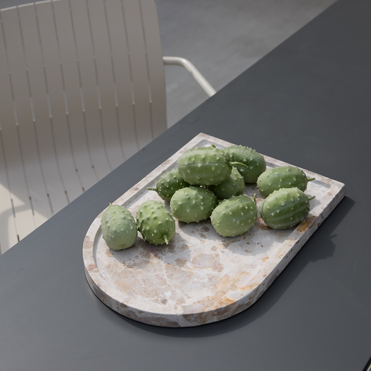 Grønne piggede frugter på et terrazzo-serveringsbræt på et mørkegråt udendørs bord med en lys stol i baggrunden.