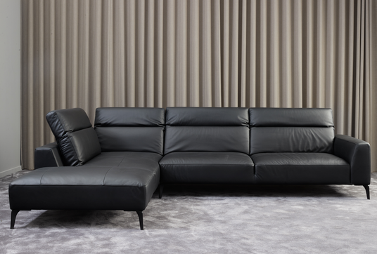 Sort læderhjørnesofa med chaiselong foran beige gardiner på gråt tæppe.
