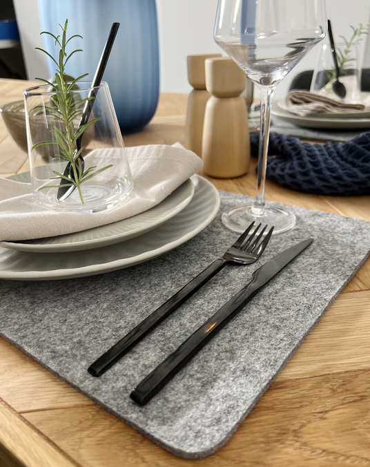 Dækket bord med gråt filt-placemat, sort bestik, stak lysegrå tallerkener, linnedserviet, vinglas og drikkeglas med en rosmarinkvist; trækværne og blå vase i baggrunden.