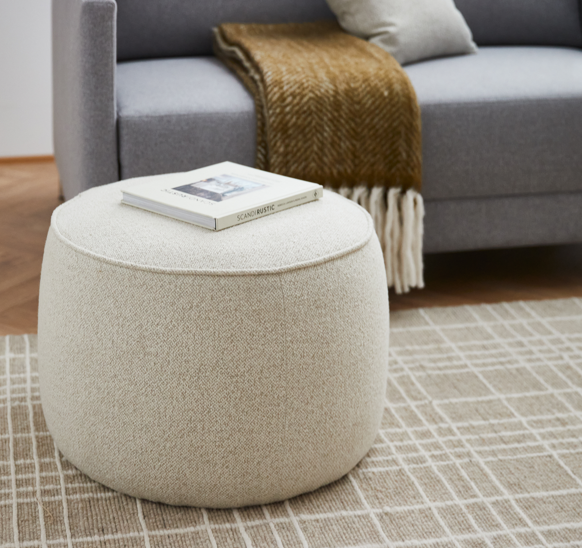 Beige rund puf på ternet tæppe med bogen Scandi Rustic ovenpå; grå sofa med pude og brun plaid i baggrunden.