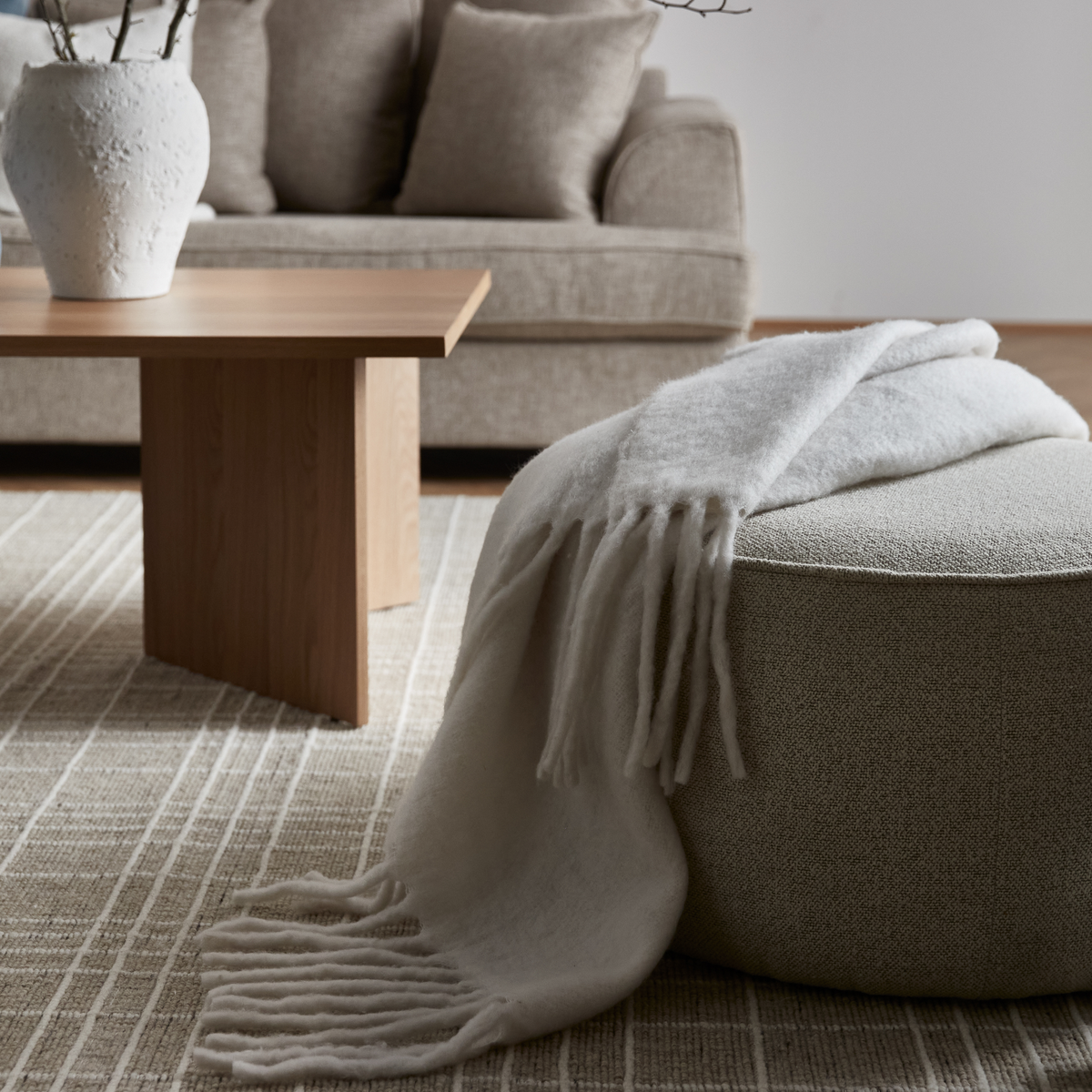 Beige stue med rund puf med lys frynset plaid, træbord med hvid vase, sofa med puder og neutral ternet tæppe.