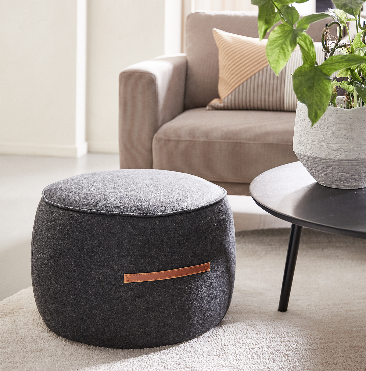 Grå stofpuf med læderhank i en neutral stue ved siden af et sort rundt sofabord med potteplante og beige sofa.