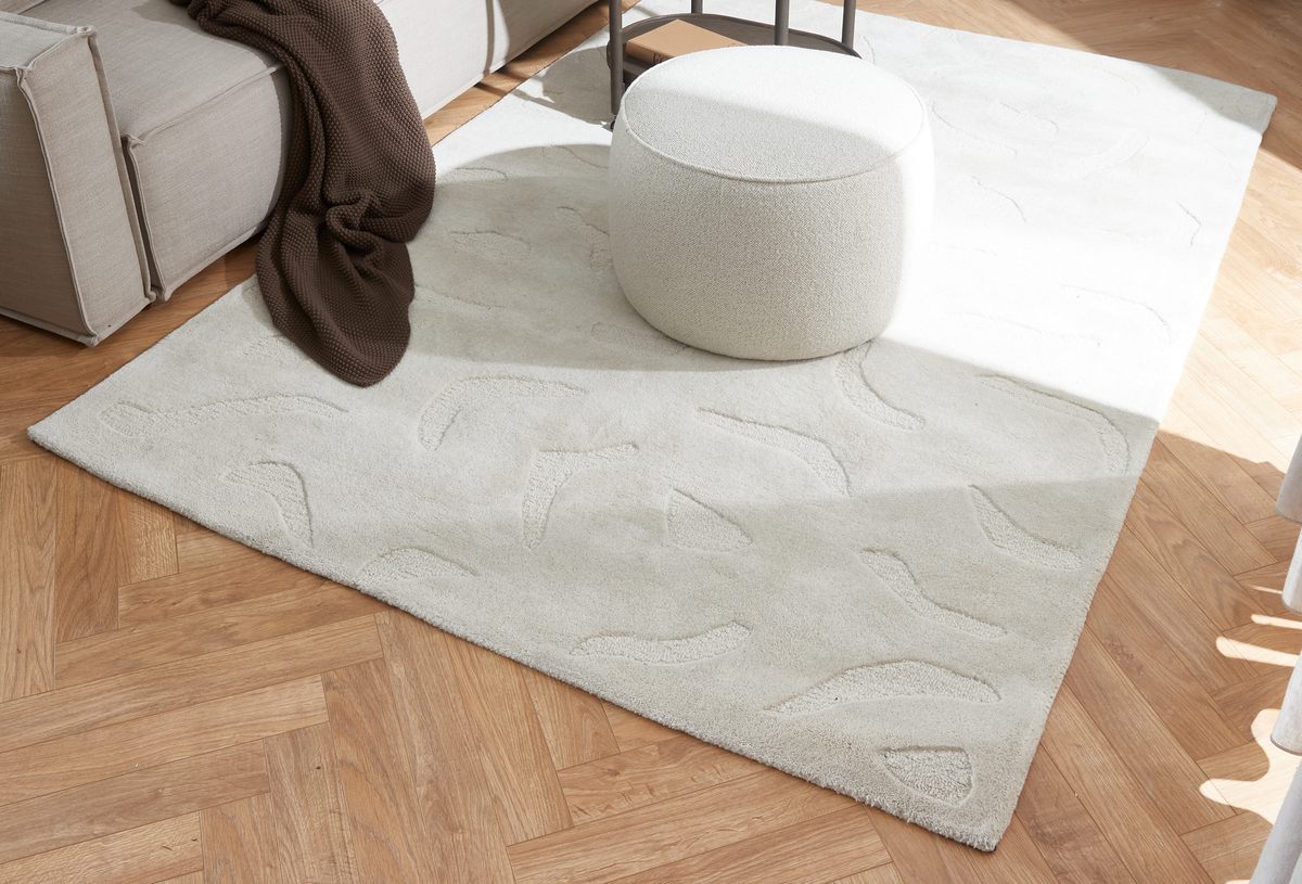 Lys beige tæppe med diskret reliefmønster på sildebensparket ved siden af en hvid puf og en beige sofa med brun plaid i dagslys.