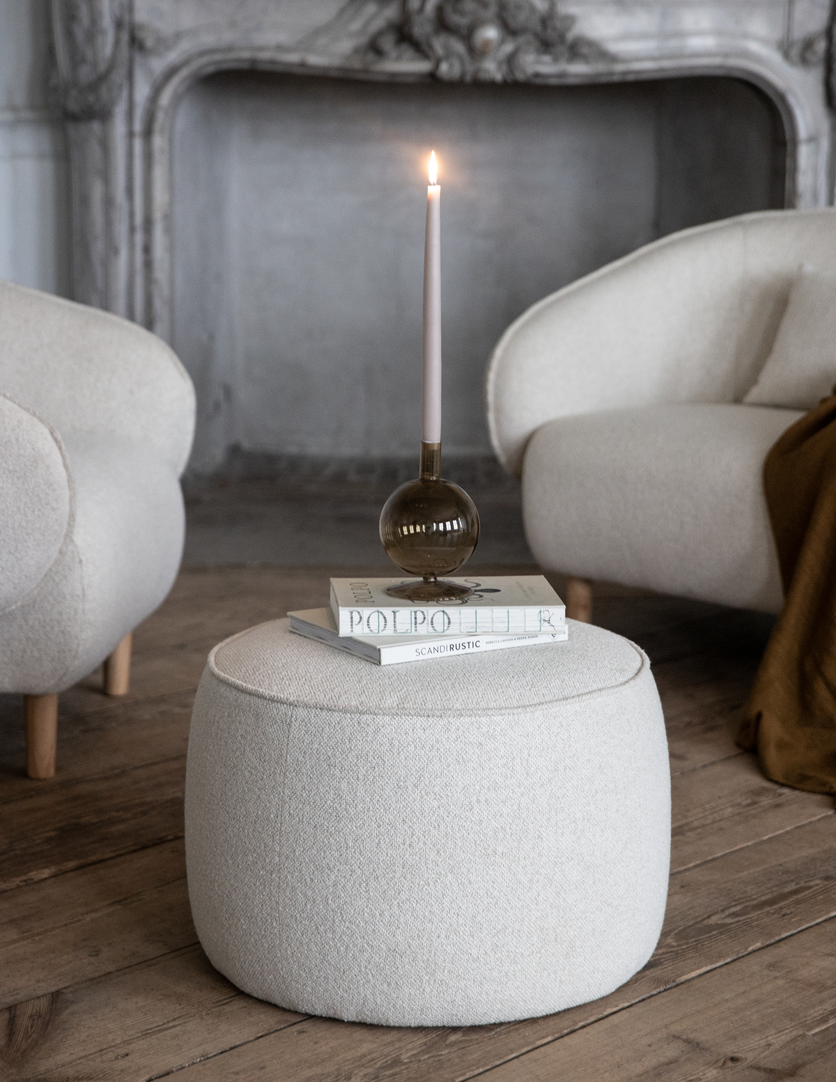 Beigefarvet bouclé-puf med bøgerne ”POLPO” og ”SCANDI RUSTIC” ovenpå, toppet med et tændt slankt lys i røgfarvet glasstage mellem to lyse lænestole ved en udsmykket pejs.