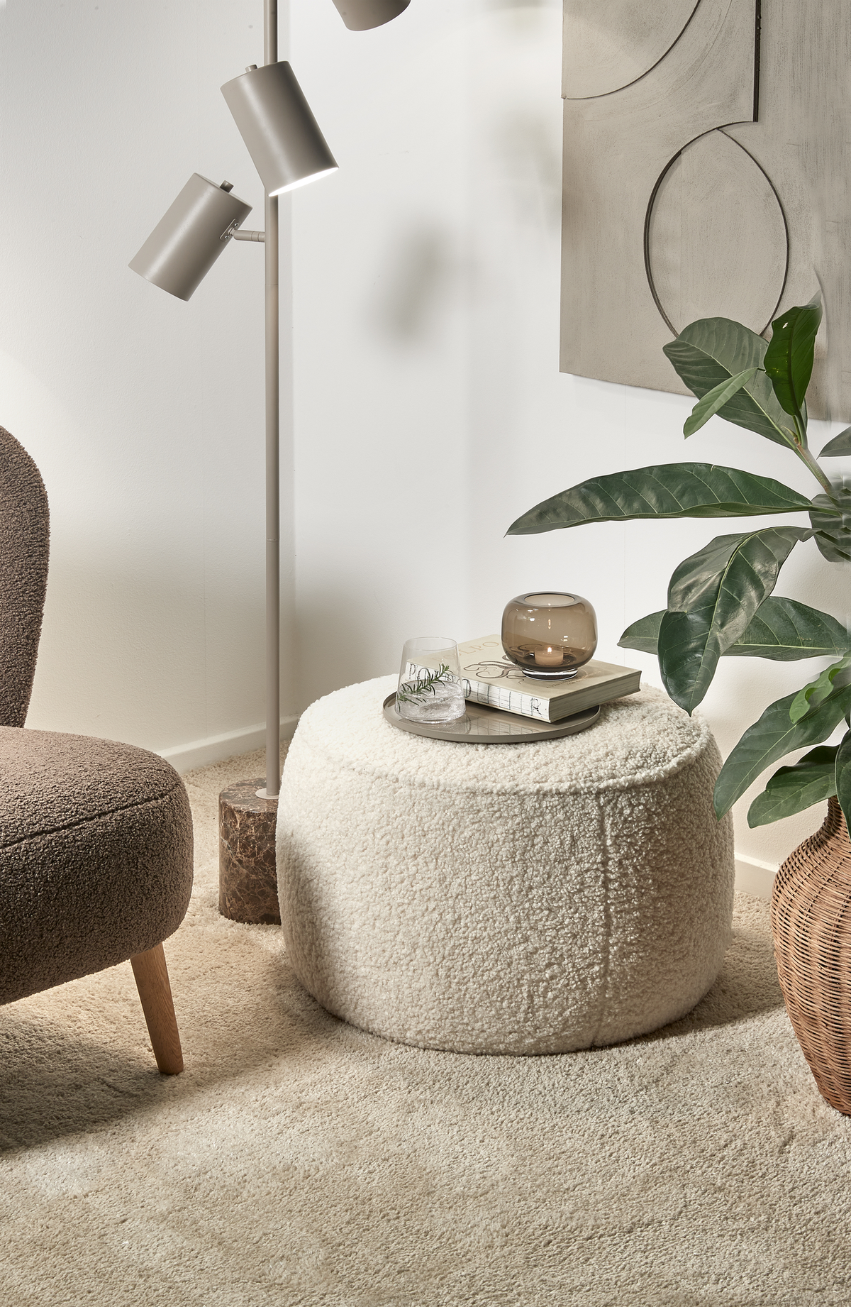 Hyggeligt hjørne i stuen med beige bouclé‑puf som sidebord med bakke, glas og lille lyslygte på, ved siden af lænestol, gulvlampe, plante i kurv og vægkunst i neutrale toner.