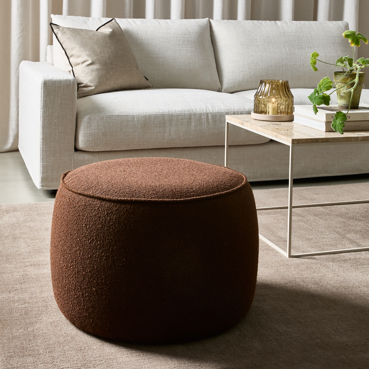 Brun rund puf i bouclé foran lys sofa og sofabord med stenplade, lysglas og grøn plante i en stilren stue.