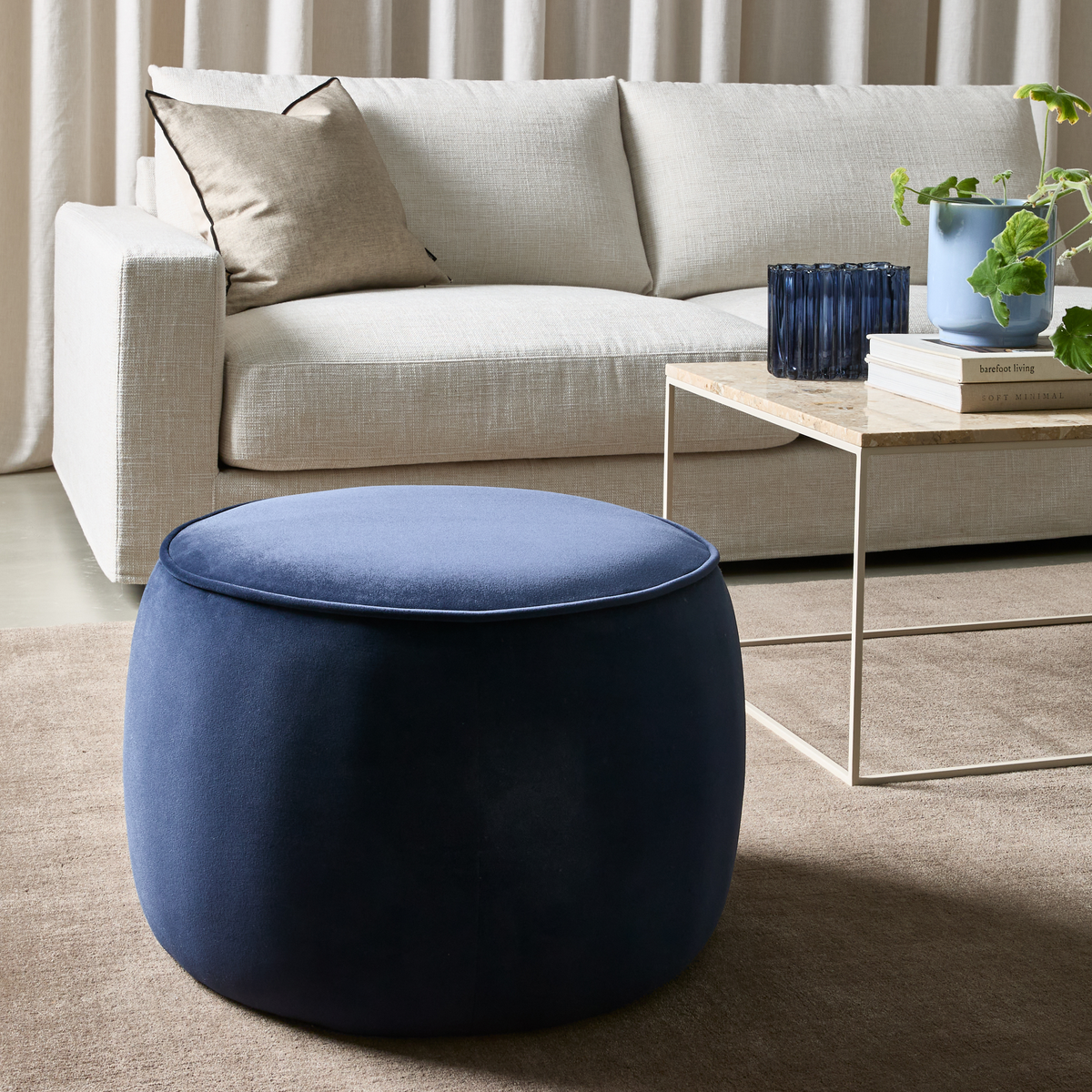 Beige sofa med pude, lyst bord med bogstakke ”barefoot living” og ”Soft Minimal” ved siden af blå glas og en plante; i forgrunden en rund mørkeblå puf på beige tæppe.