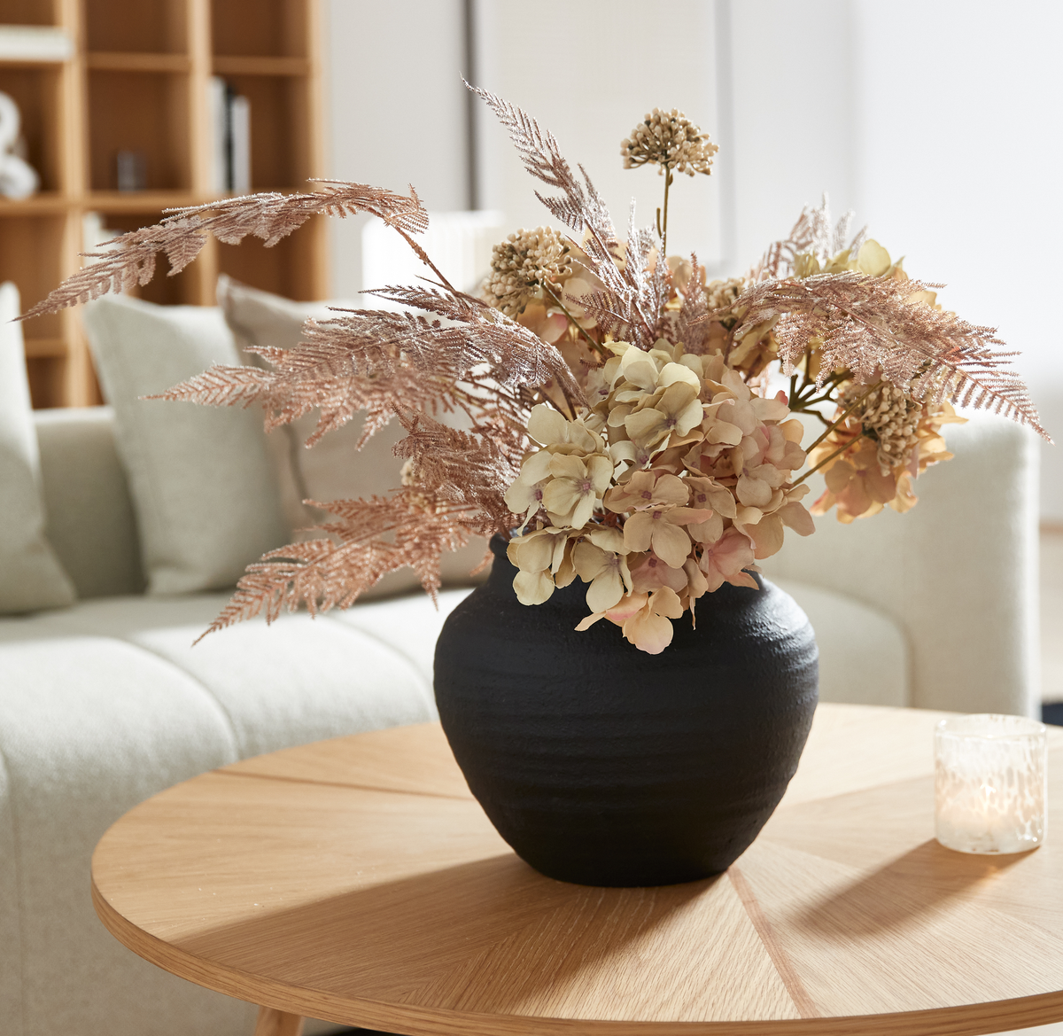 Sort vase med tørrede blomster på rundt sofabord i lys skandinavisk stue, med et lille fyrfadslys ved siden af.