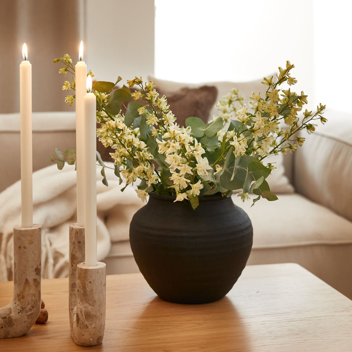 Sort vase med hvide blomster og eucalyptus på et træbord ved siden af to tændte stearinlys i stager af sten foran en beige sofa.