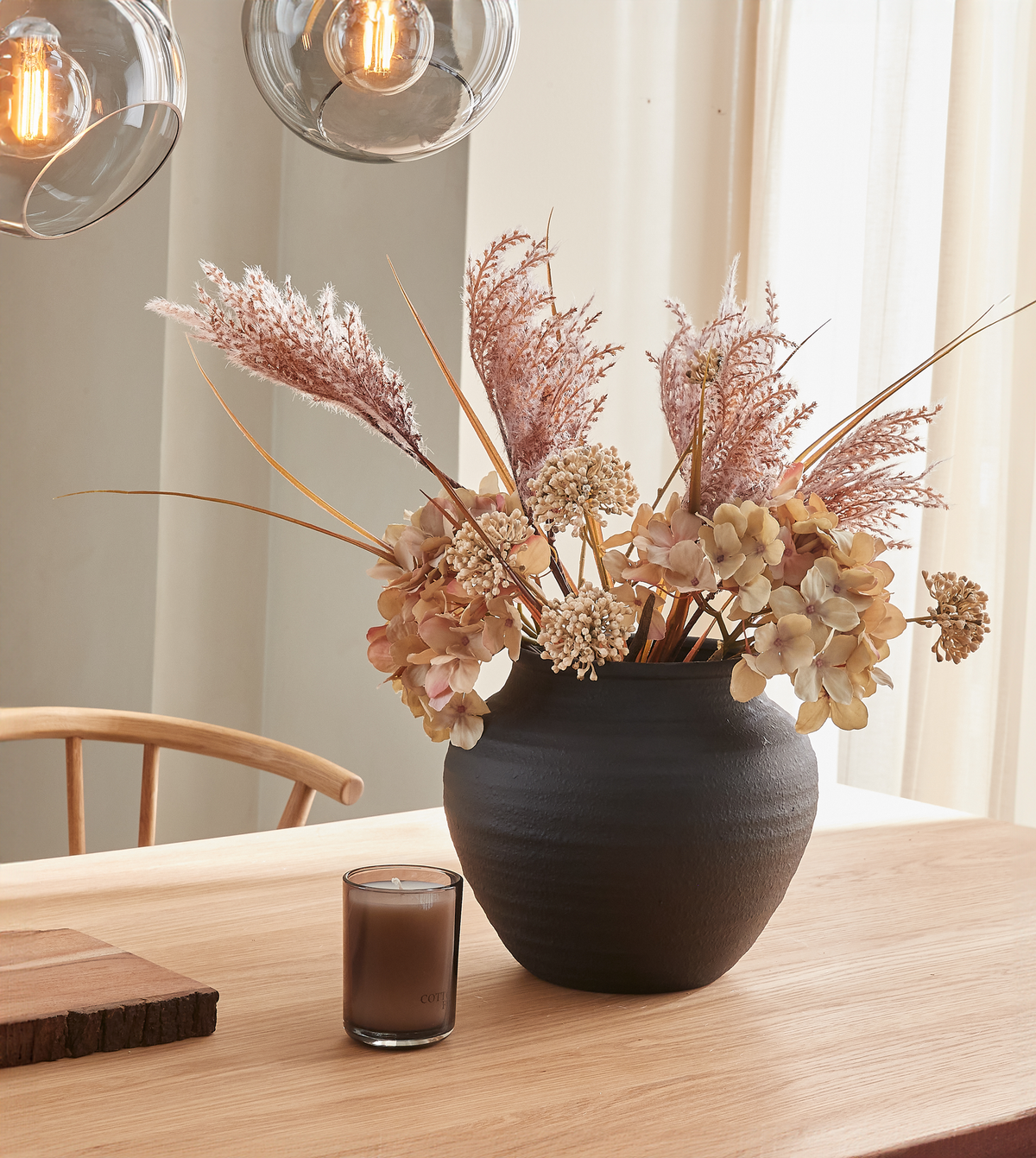 Sort keramisk vase med tørrede blomster på et lyst træbord, et lille brunt lys ved siden af og varme pendellamper over.
