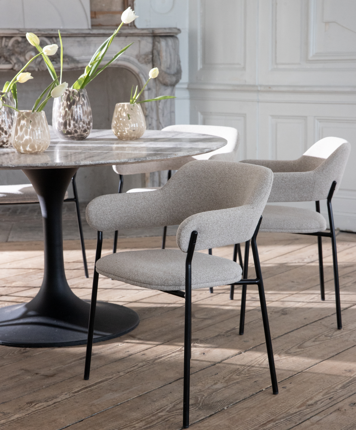 Lyst spisestue med rundt marmorbord på sort sokkel, lyse beige polstrede spisestole med sorte ben og glasvaser med hvide tulipaner på bordet.
