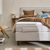 Lyst soveværelse med polstret seng i grå tekstil, brunt sengetøj og puder, blåternet pude, væghylde i træ med lampe og bøger, fletstol og beige-hvidternet tæppe.