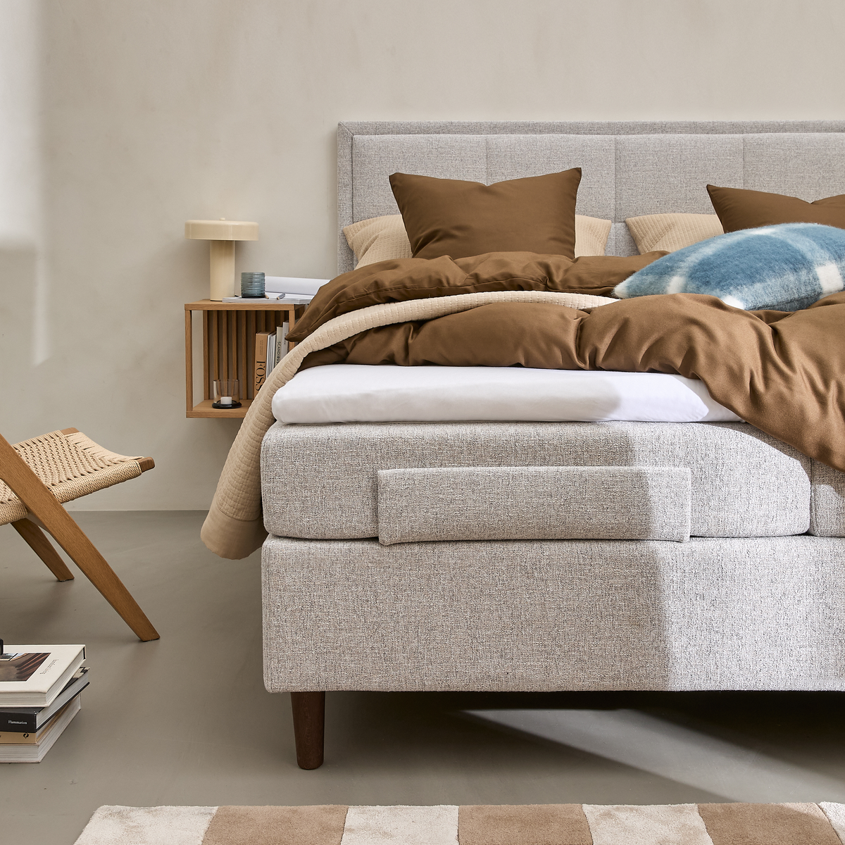 Lyst soveværelse med polstret seng i grå tekstil, brunt sengetøj og puder, blåternet pude, væghylde i træ med lampe og bøger, fletstol og beige-hvidternet tæppe.