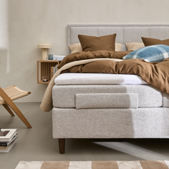 Lyst soveværelse med polstret seng i grå tekstil, brunt sengetøj og puder, blåternet pude, væghylde i træ med lampe og bøger, fletstol og beige-hvidternet tæppe.