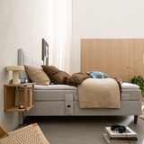 Lyst og minimalistisk soveværelse med en grå polstret seng med beige og brunt sengetøj, en væghylde med bordlampe og bøger, skabe i træ, en grøn plante og en bogstak med hovedtelefoner, hvor øverste bog hedder “New Nordic Houses”.