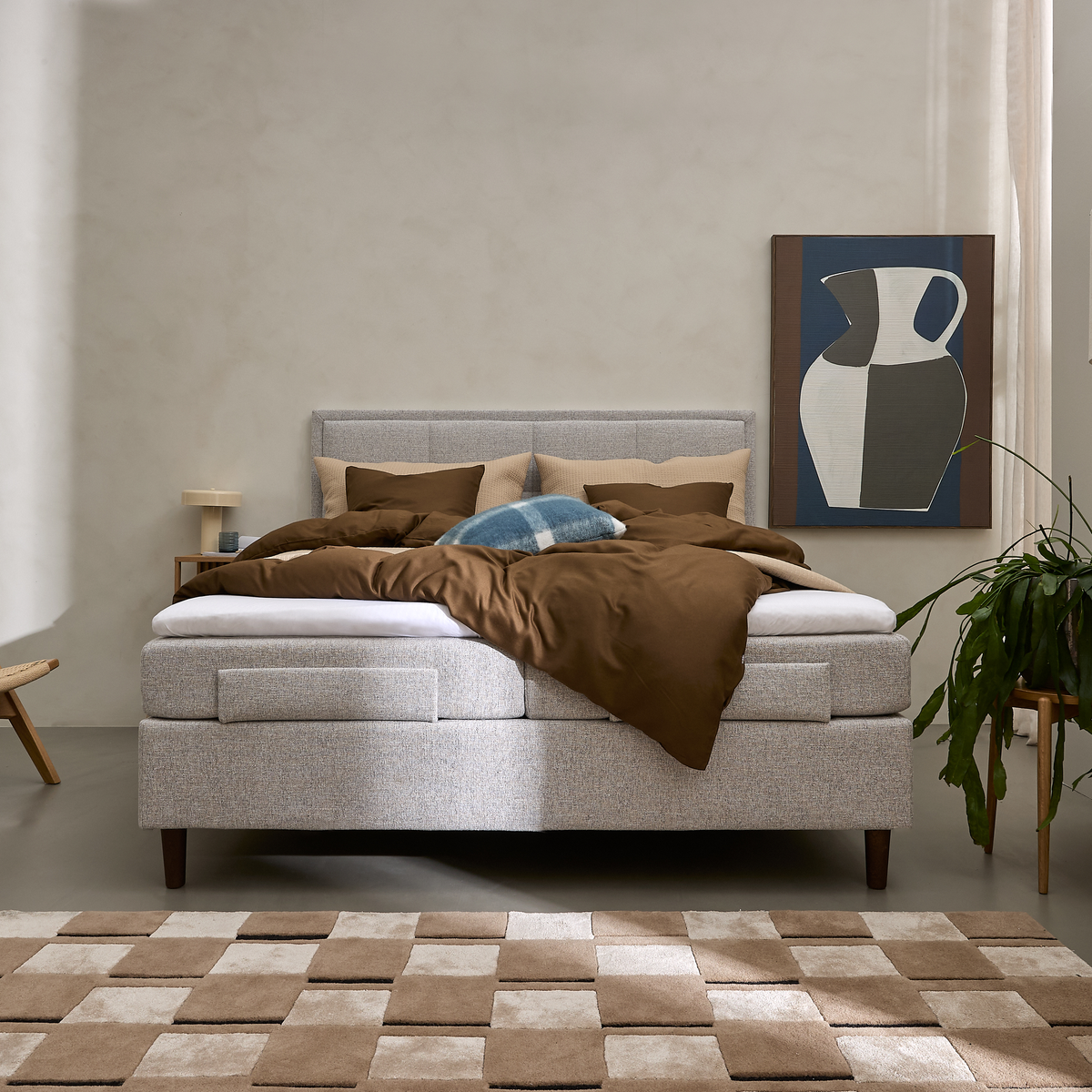 Grå kontinentalseng med brunt sengetøj, blå pyntepude, kunst med vase, grøn plante og beige‑brunt ternet tæppe i et lyst soveværelse.