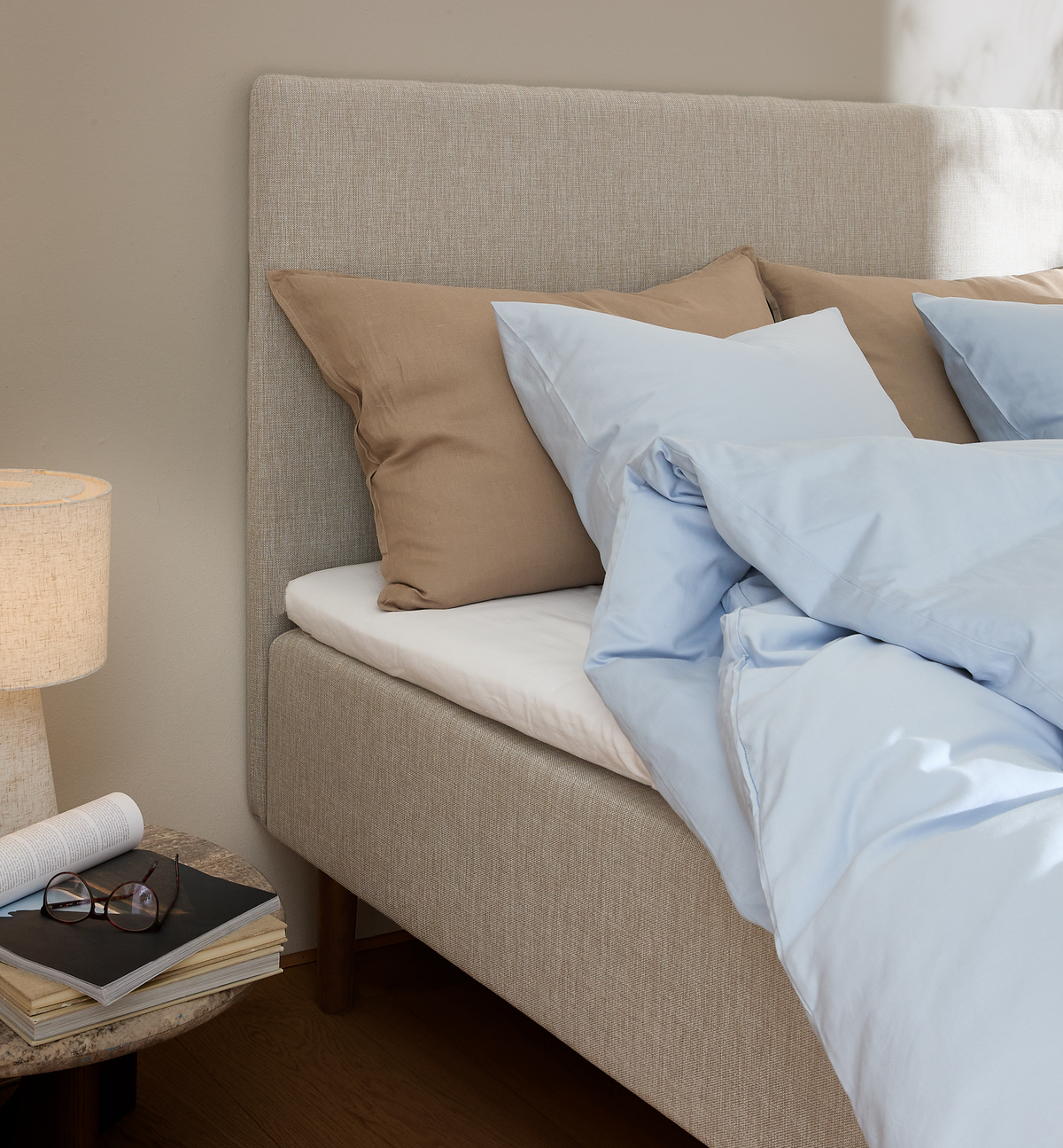 Beige seng med lyseblåt sengetøj og brune puder ved siden af et sengebord med lampe, bøger og briller i blødt dagslys.
