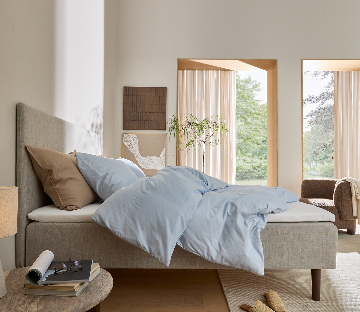 Roligt soveværelse med seng i beige og lyseblåt dynebetræk, store vinduer med lyse gardiner, plante, lænestol og sengebord med lampe og bøger i blødt dagslys.