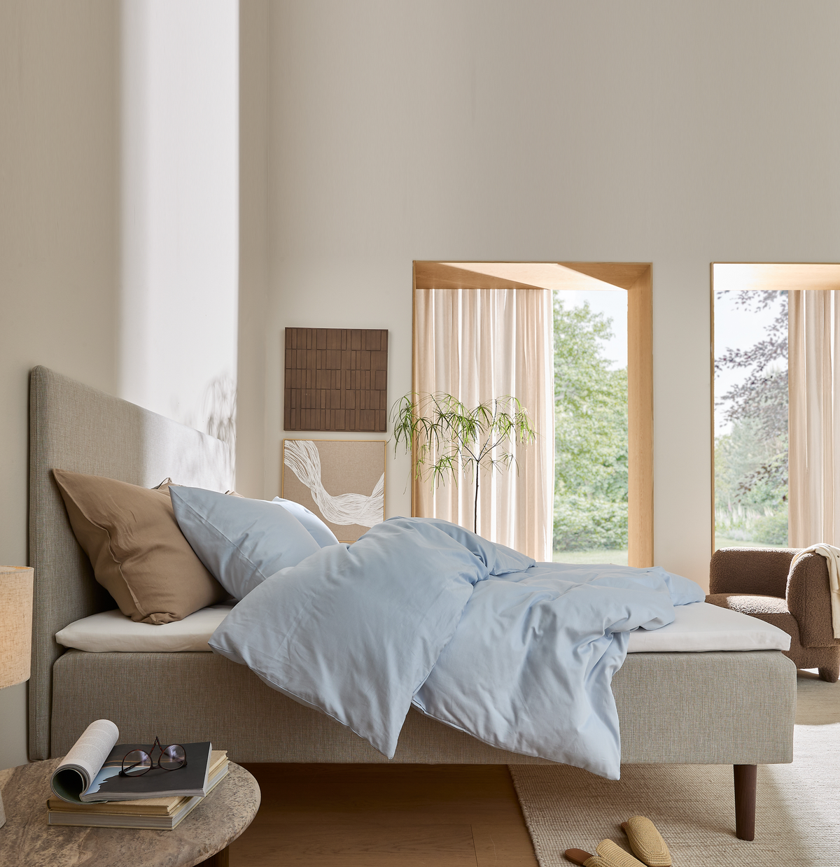 Lyst soveværelse med beige polstret seng, lyseblåt dynebetræk, neutrale puder, lyse gardiner og store vinduer mod grøn natur; sengebord med bøger og briller.