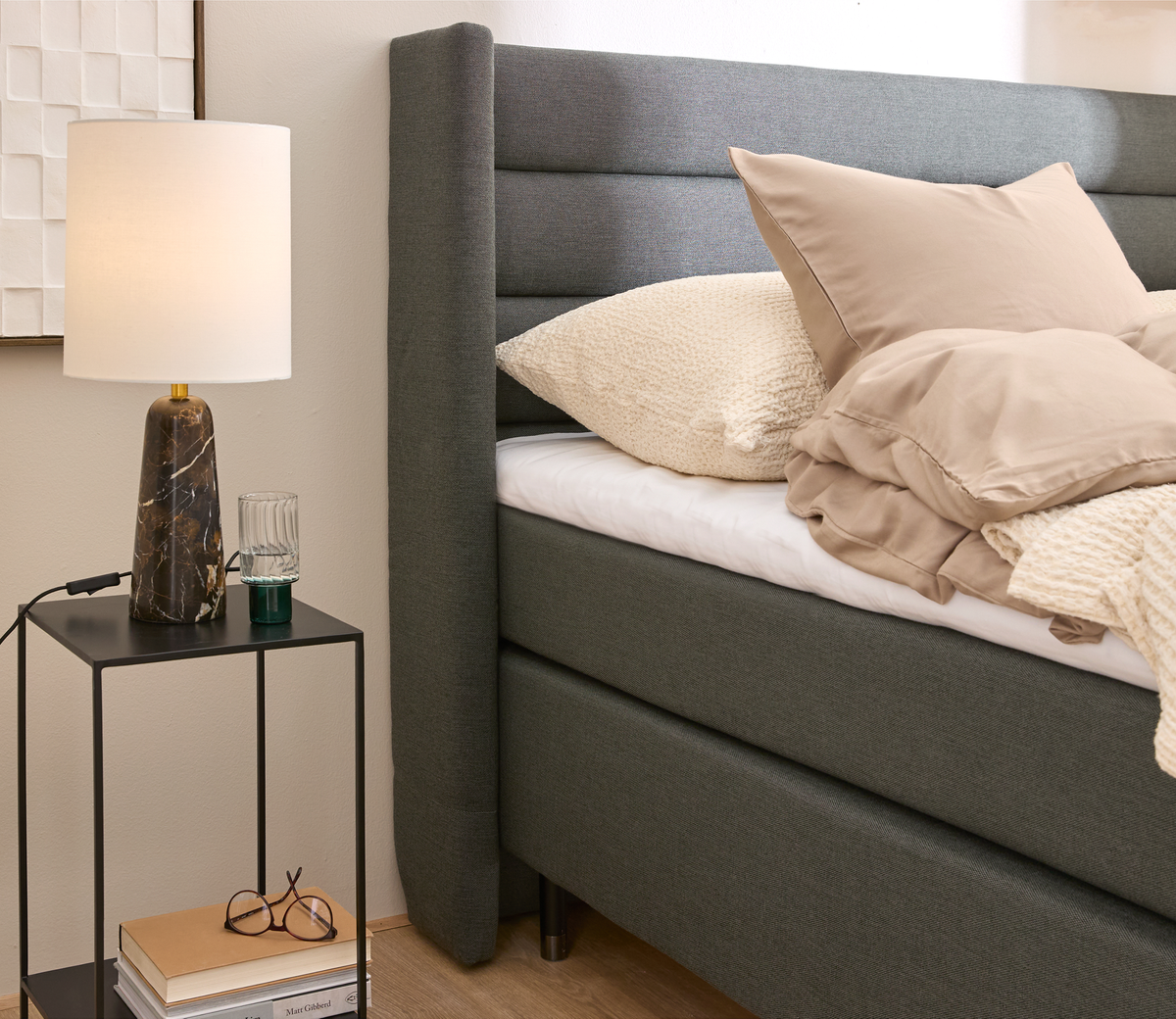 Soveværelse med grå polstret seng, beige sengetøj og natbord med marmorbordlampe, glas og bøger.
