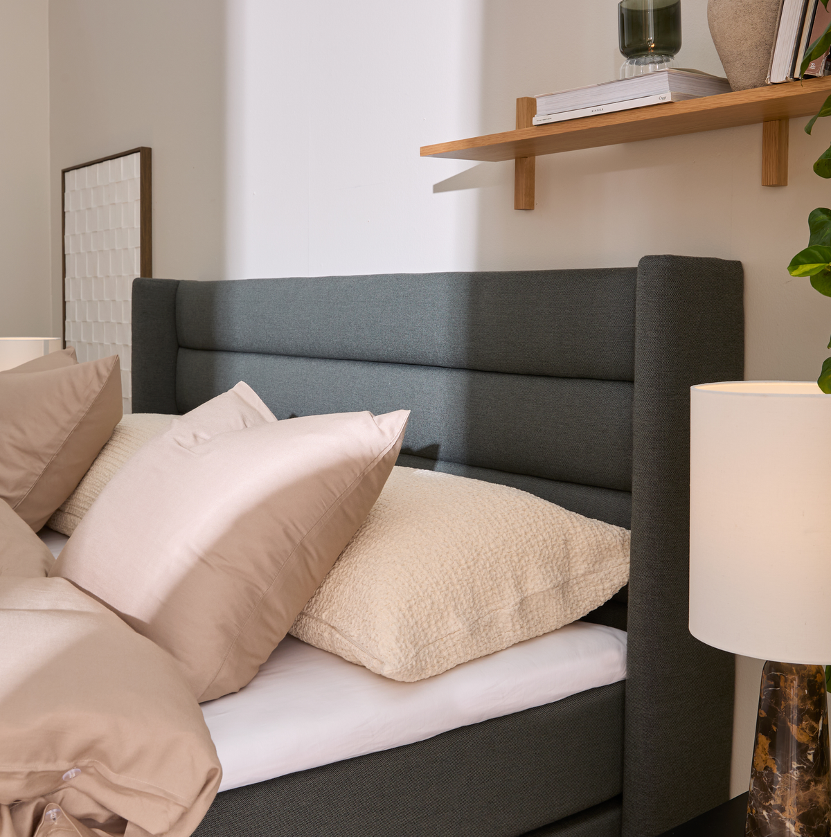 Nærbillede af seng med grå polstret sengegavl, beige puder og bordlampe; træhylde med bøger og vase i blødt dagslys.