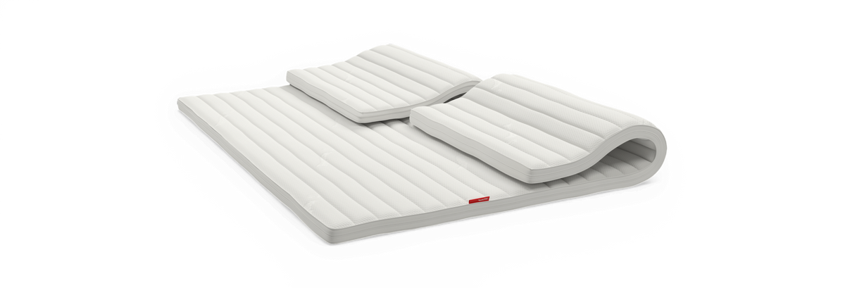 Hvid ribbet topmadras med to matchende ergonomiske puder, rullet i den ene ende, fritlagt mod sort baggrund.