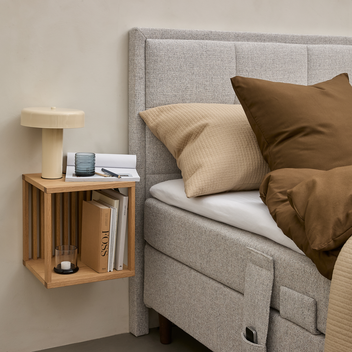 Lys grå polstret seng med beige og brunt sengetøj ved siden af et væghængt natbord i eg med cremefarvet lampe, glas, notesbog, pen, fyrfadslys og bøger, bl.a. FOSS.