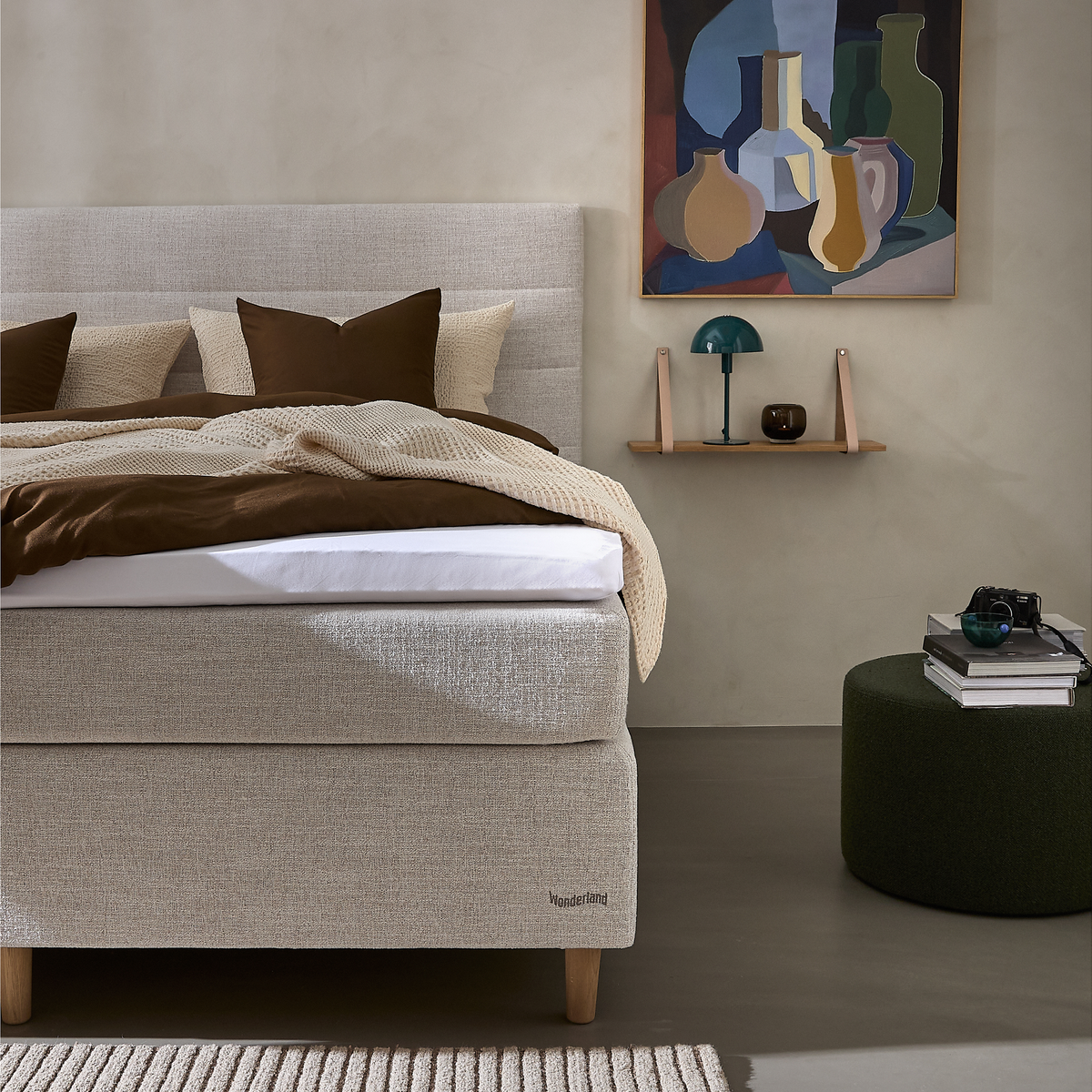 Beige polstret seng med brunt og cremefarvet sengetøj, mærket Wonderland, væghylde med grøn bordlampe og kunst over; mørkegrøn puff med bøger og kamera i et roligt soveværelse.