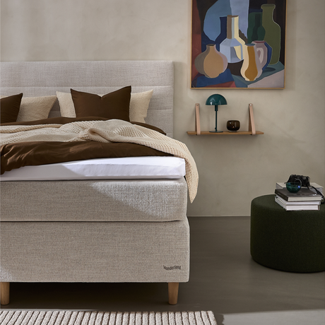 Beige polstret seng med brunt og cremefarvet sengetøj, mærket Wonderland, væghylde med grøn bordlampe og kunst over; mørkegrøn puff med bøger og kamera i et roligt soveværelse.