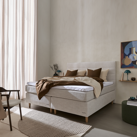 Lyst soveværelse med beige kontinentalseng på træben, brun- og beige sengetøj, høje lyse gardiner, neutral vævet tæppe og væghylde med små lamper og kunst.