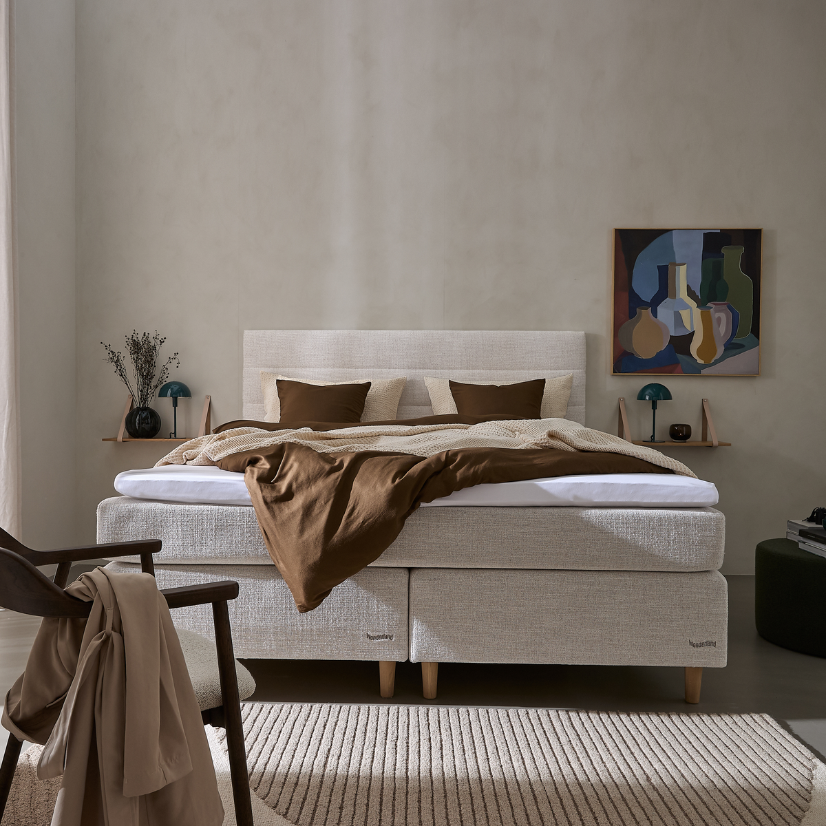 Lyst soveværelse med beige kontinentalseng, brunt sengetøj og strikket plaid, to små væghylder med turkise bordlamper og farverigt maleri, stol med beige kåbe og stribet tæppe.