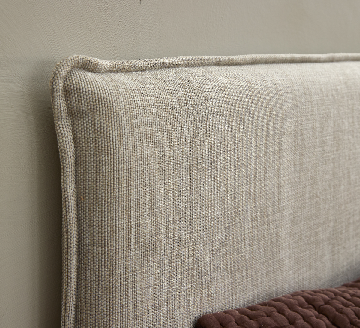 Nærbillede af beige polstret sengegavl med pipingsøm; brun vatteret quilt ses nederst mod en neutral væg.