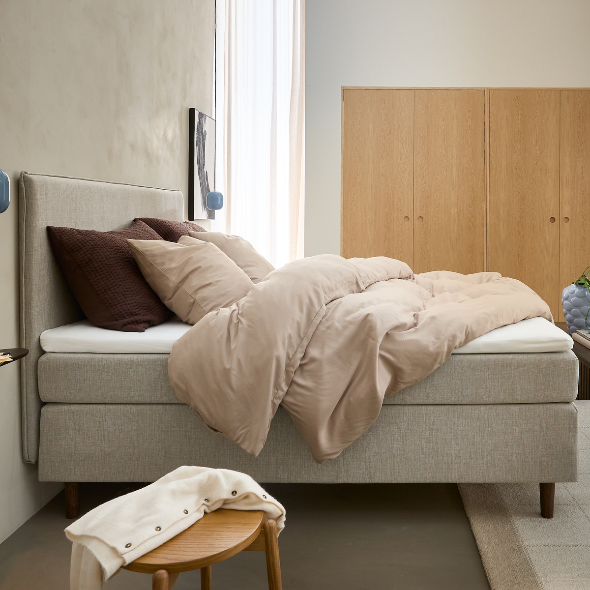 Lyst soveværelse med beige sengetøj på en polstret seng, brune puder, lys træ-garderobe og en skammel med en hvid trøje.