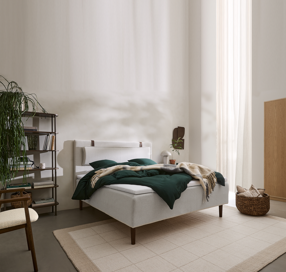 Lyst soveværelse med lysegrå polstret seng, mørkegrønt sengetøj og beige plaid, stor lys tæppe, åben reol med bøger og planter, flettet kurv med puder og lette gardiner i dagslys.