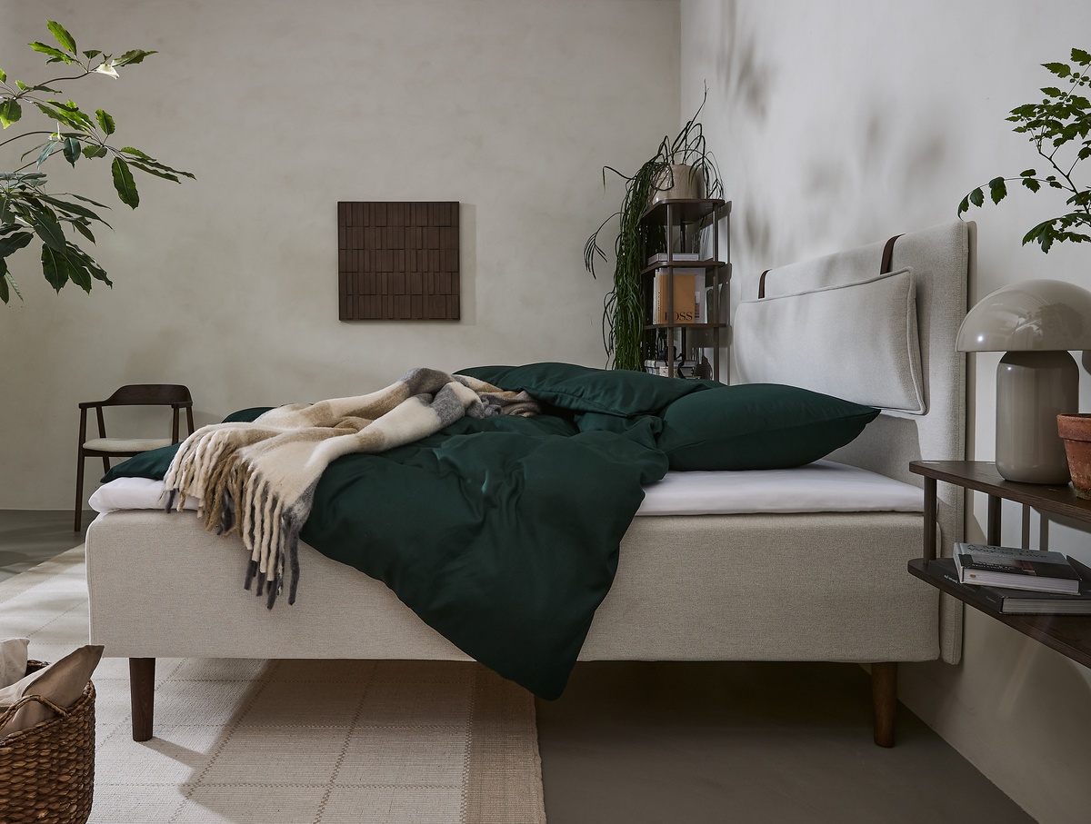 Lyst soveværelse med beige polstret seng, mørkegrønt sengetøj, uldtæppe og planter.