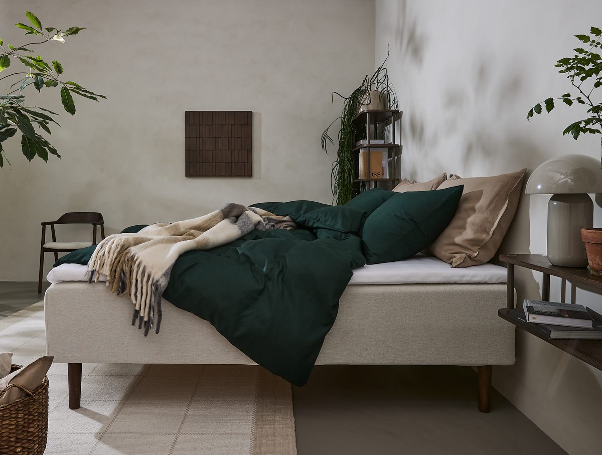 Soveværelse med beige polstret seng, mørkegrønt sengetøj, sandfarvede puder og en uldtæppe, med planter, reol og bordlampe i blødt dagslys.