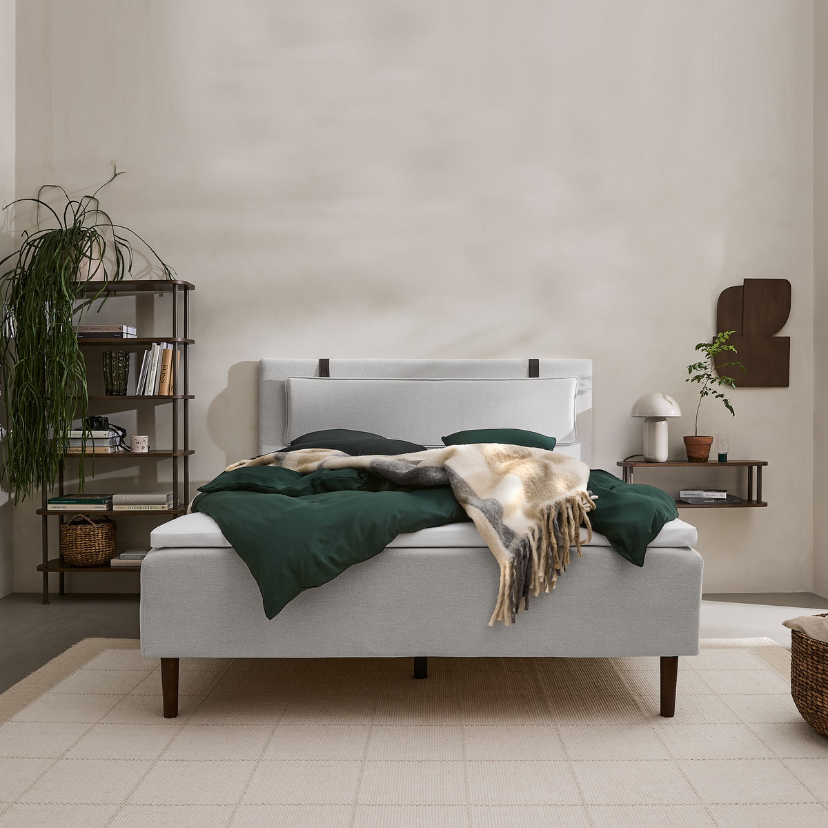 Lyst soveværelse i skandinavisk stil med lysegrå polstret seng, mørkegrønt sengetøj og frynset plaid, hylder med bøger og planter samt beige tæppe.