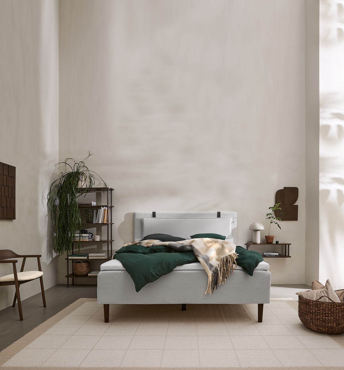 Lyst, minimalistisk soveværelse med lysegrå seng, grønne sengesæt og beige plaid; reol og plante ved siden, bordlampe og kurv på et beige tæppe.