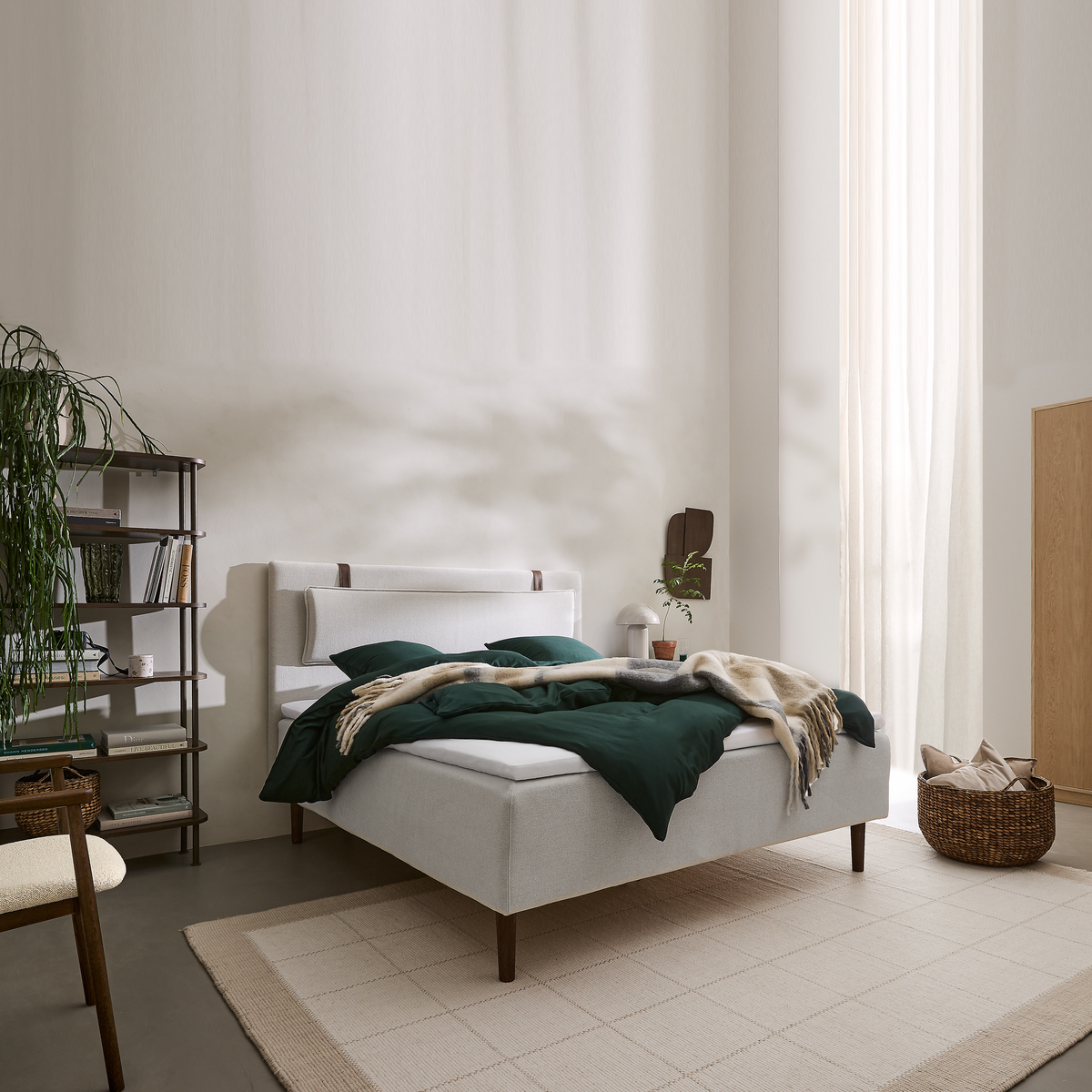 Lyst soveværelse med hvidt polstret seng, mørkegrent dyne og beige plaid, lyse gardiner, reol med planter og en flettet kurv med puder på et lyst tæppe.
