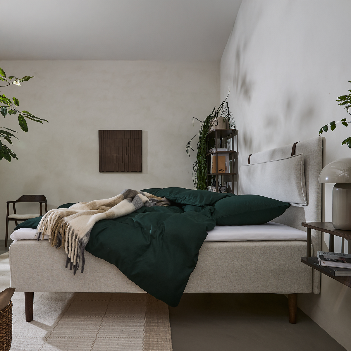 Soveværelse med beige seng, mørkegrønt sengetøj og plaid, planter og reol i hjørnet.