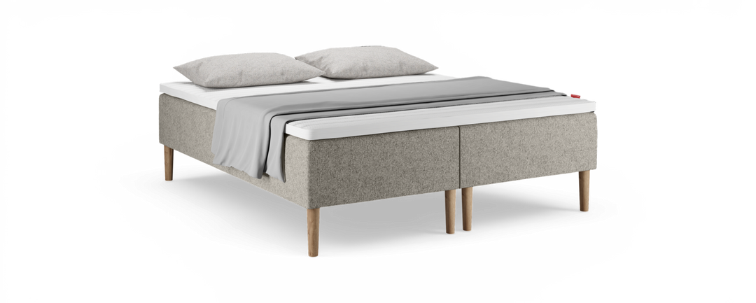 Beige polstret seng med træben, hvid topmadras, to grå puder og et lysgråt plaid over sengen.