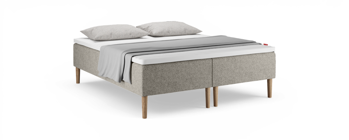 Beige polstret seng med træben, hvid topmadras, to grå puder og et lysgråt plaid over sengen.