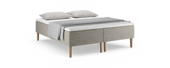 Beige polstret seng med træben, hvid topmadras, to grå puder og et lysgråt plaid over sengen.