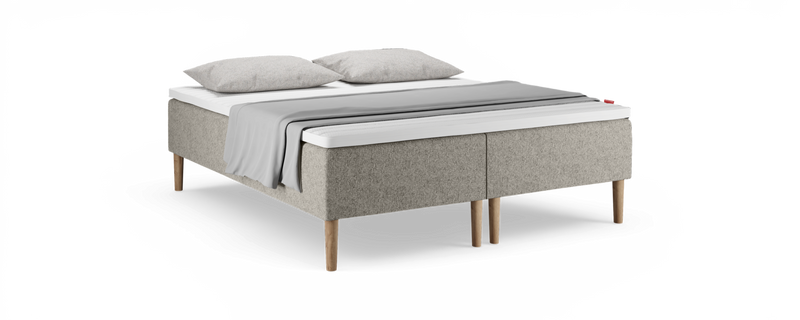Beige polstret seng med træben, hvid topmadras, to grå puder og et lysgråt plaid over sengen._ps