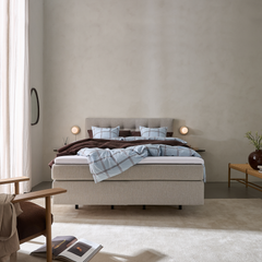 Lyst soveværelse med beige polstret seng, blåternet sengetøj og brun plaid; væglamper, gardiner og stor tæppe i blødt dagslys.